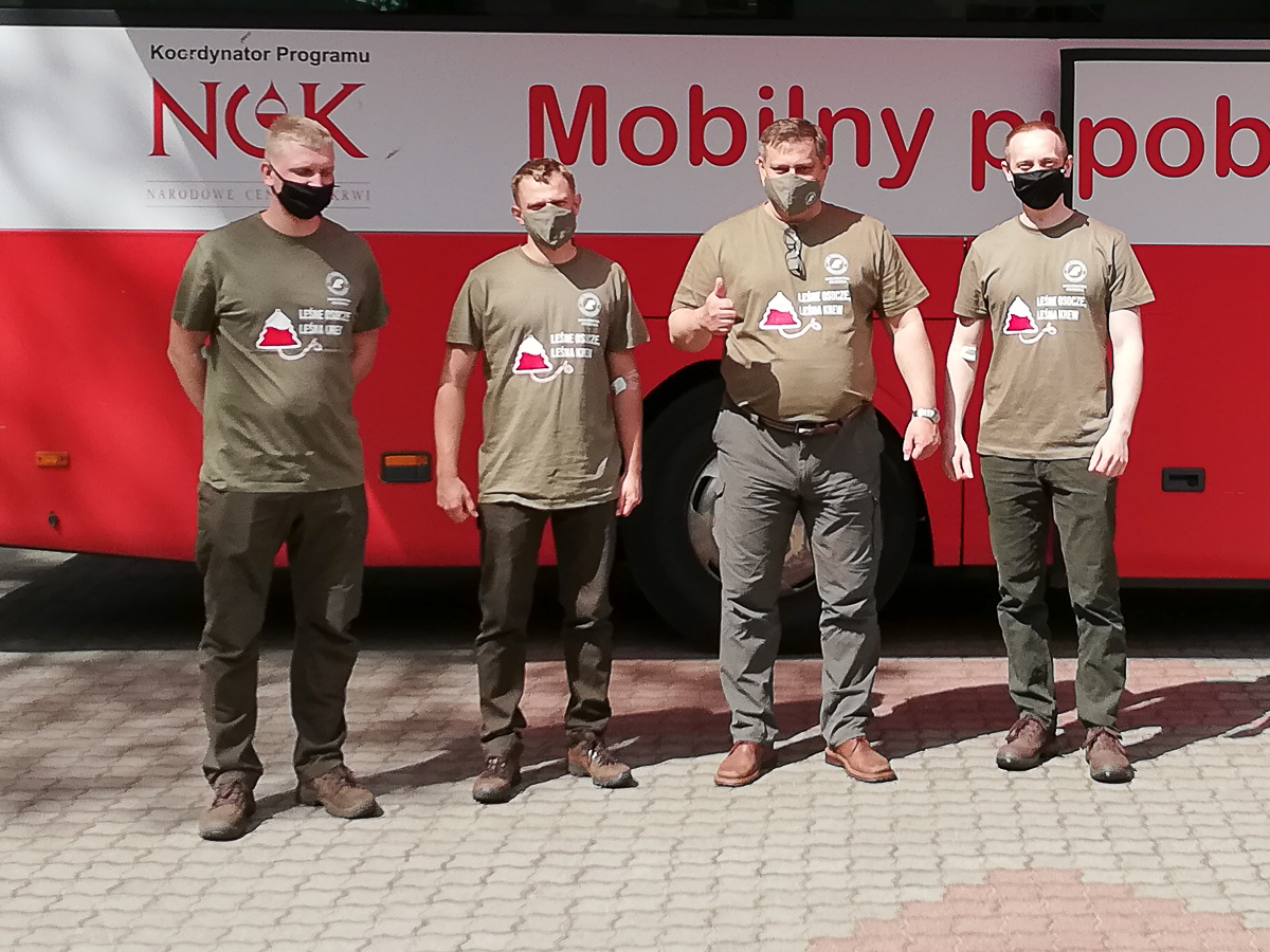 Czterej mężczyźni stoją ubrani w okolicznościowe koszulki z napisem leśne osocze leśna krew, w tle autobus w kolorze biało-czerwonym.